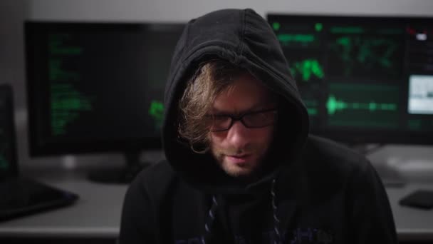 Κοντινό πλάνο ενός χάκερ που με κουκούλα με γυαλιά που είναι ντυμένος με μαύρα ρούχα που κάθεται σε μια καρέκλα στο δωμάτιο και Βάλε χρήματα για hacking δικτύου και αποκτά πρόσβαση σε σημαντικές πληροφορίες. — Αρχείο Βίντεο