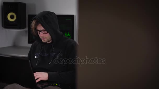 Pleased Hacker en una sudadera con capucha negra con color kayushonom sentado en su natbukom y comprometido en programas de hacking. En el fondo hay unas cuantas pantallas de computadora, en las que puede rastrear satélites — Vídeos de Stock