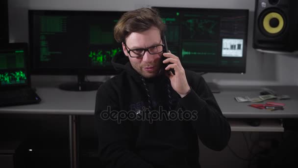 Der Mann im Computerraum. Junger Mann mit Brille sitzt auf einem Stuhl und spricht mit seinem fokussierten Smartphone. auf Monitoren Prozess des Hacking. gegen ausgerüstete Computerstation. — Stockvideo