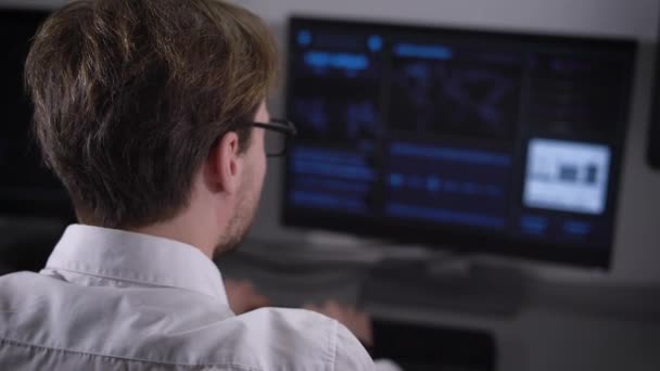 Experienced IT specialist works on a new program on two computers. Young man in glasses and white shirt writes the source code of the software. — Stock Video