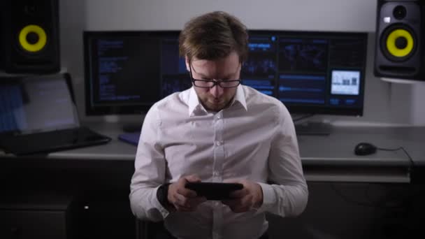 El joven desarrollador de juegos en camisa blanca y pantalones negros está sentado en la oficina con la tableta en sus manos y probando la nueva versión del producto. Freelancer masculino jugando durante la hora del almuerzo . — Vídeos de Stock