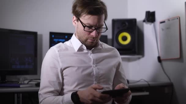 Büroangestellte hält ein Smartphone in der Hand und spielt ein Handyspiel. Es konzentriert sich auf den Prozess. Ein Mann mit Brille trägt ein weißes Hemd, seine Hand Smartwatches. im Hintergrund ist mehr als ein Monitor — Stockvideo