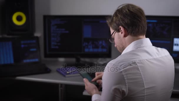 Närbild av ung frilansare klädd i vit skjorta och med smartwatch på handen arbetar på Pc på hans rum hemma. Programmeraren sitter vid bordet, att skriva på tangentbordet och använder sin mobiltelefon. — Stockvideo