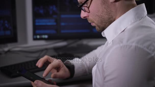 Adult businessman with glasses and white shirt sitting in his office and engaged in view of popular magazines about the finances on your phone, and then he looks at the web page on your laptop. — Stock Video