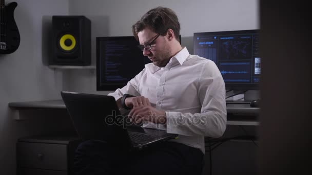Mladý programátor v bílé košili a stylové brýle, pracují na notebooku, který je na kolenou na pozadí několika monitorů s více informací. — Stock video