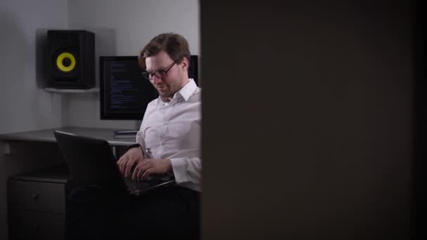 Porträt eines jungen Mannes, der am Laptop arbeitet und sein Gadget mit Smartwatch auf der Hand hält. Entwickler zeitgenössischer Armbanduhren sitzt vor Computermonitoren und betrachtet seine Erfindung. — Stockvideo