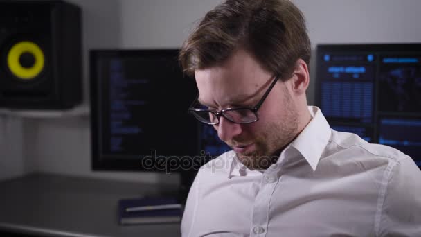 Hombre serio con gafas, vestido con una camisa blanca sentado en la zona de trabajo e imprime un informe en su computadora portátil. El hombre se centró en su trabajo. En el fondo hay varias pantallas de monitor — Vídeo de stock