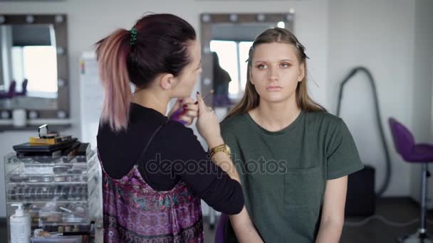 Proceso de trabajo de maquillaje de aplicación en el estudio de belleza. Experto profesional está fijando polvo en la cara de las niñas. Rubia joven modelo está sentado mientras que el profesor profesional está demostrando la técnica moderna . — Vídeos de Stock