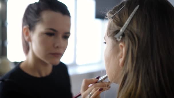 Επαγγελματία make-up artist στη βιομηχανία της μόδας κάνει μακιγιάζ νεαρό μοντέλο που κάθεται σε μια καρέκλα στο ατελιέ της ομορφιάς. Κορίτσι με ένα πινέλο στα χέρια του θολή στο φόντο. — Αρχείο Βίντεο