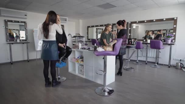 Dos artistas de maquillaje principiantes están capacitados para aplicar cosméticos profesionales de maquillaje. Como modelos tienen dos chicas, una rubia, la otra con el pelo oscuro. En el fondo, una gran cantidad de espejos, donde la mirada del modelo — Vídeo de stock