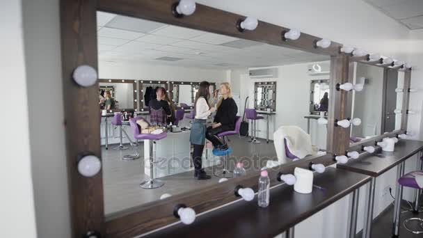 La vista desde el espejo con luces, que se encuentra en un vestidor de luz. Dos maquilladores maquillando a una joven modelo, que en silencio esperan su finalización en esta etapa y comienzan a trabajar con el cabello — Vídeos de Stock