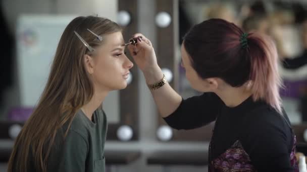 Makeupartist betonar formen på ögonbrynen modell med brun ögonbrynsgel. Modellen sitter tyst. Hennes ansikte har redan gjorts fuktkräm, foundation, concealer, eyeliner, svart mascara — Stockvideo