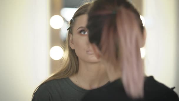 Primer plano de la joven portada profesional durante el proceso de maquillaje en el salón de belleza. Visagiste joven aplicando sombra de ojos en la cara de los modelos. Artista morena creando una imagen de modelo para fotografía futura . — Vídeo de stock