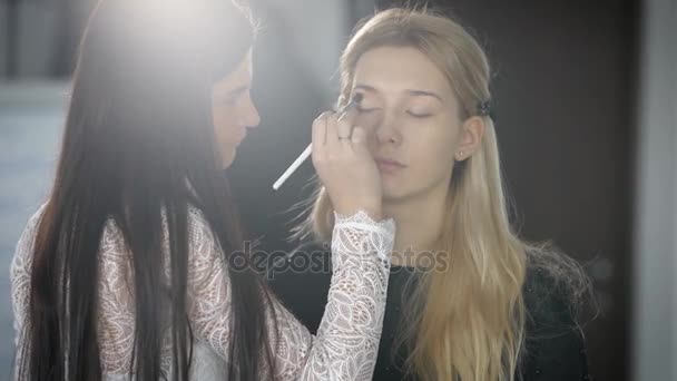 Hermosa rubia con cejas delgadas, pelo largo espera el maquillaje. Maquillaje artista hace un maquillaje profesional, ahora está en la etapa de aplicar sombra de ojos en los párpados cerrados a la joven — Vídeo de stock