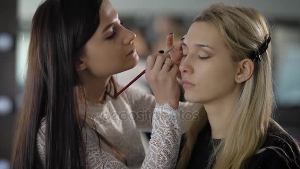 Unga makeupartist applicerar ögonskugga på framsidan en ung modell med tunna ögonbryn. Den modell blond lång och välpreparerade hår, som tas bort från ansiktet med hjälp av plast clips — Stockvideo