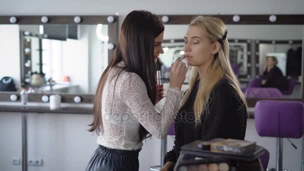 Nahaufnahme des Make-up-Prozesses im stylischen Schönheitssalon. Die junge Visagistin konturiert die Mundlinie mit speziellen Lippenstiften auf dem Gesicht der Models. Brünette Künstlerin schafft ein Bild des Modells für zukünftige Fotografie. — Stockvideo