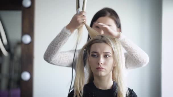 Un institut de beauté. Ferme là. Le client se prépare pour une fête à la mode. Le coiffeur avec l'aide d'une ondulation du fer à friser fait volume sur la tête . — Video