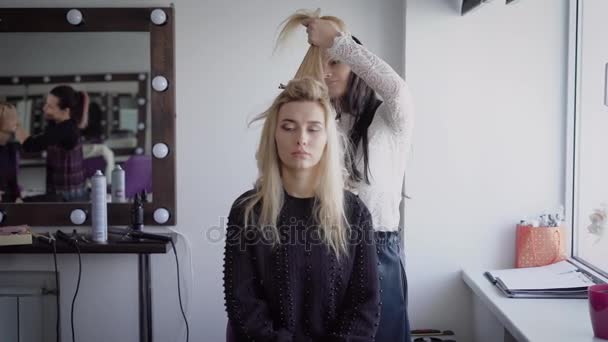 Joven peluquera femenina de salón de belleza de lujo está de pie y trabajando con la joven modelo rubia con su plancha. Hermosa mujer rubia está sentada en la silla y esperando el final del proceso . — Vídeo de stock