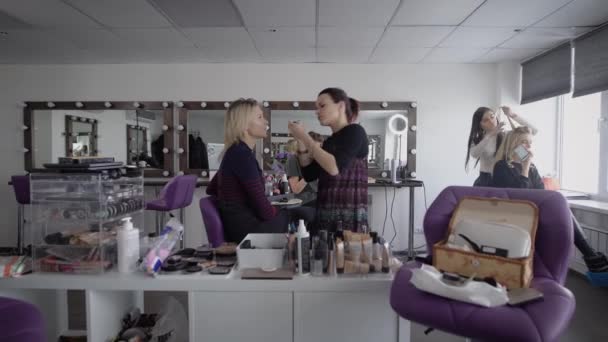 Schoonheidssalon. De master in een make-up traint drie vriendinnen naar de finale op de Universiteit. Het meisje is van toepassing met een borstel van de schaduw op de top. De make-up artiest doet professioneel een make-up. — Stockvideo