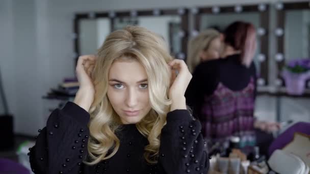 Nahaufnahme einer jungen schönen Frau im Luxus-Schönheitssalon, nachdem sie ihre Frisur mit dem Make-up-Experten im Hintergrund beendet hat. Dame mit langen Locken sitzt und glättet ihr Haar und schaut an — Stockvideo