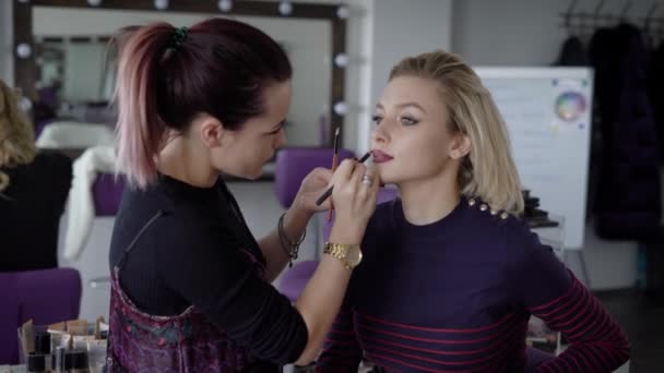 Il padrone in un trucco prepara il modello a esposizione in istituto di bellezza. La ragazza si rivolge con una matita il rossetto scuro per labbra su un labbro superiore. L'artista di trucco professionalmente fa un trucco . — Video Stock