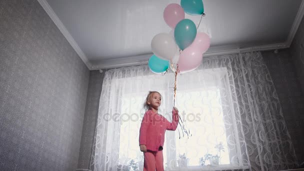 Little girl with blond hair, dressed in a pink blouse and pants jumping on the bed, which stands in front of the window and holding colorful balloons in hands — Stock Video