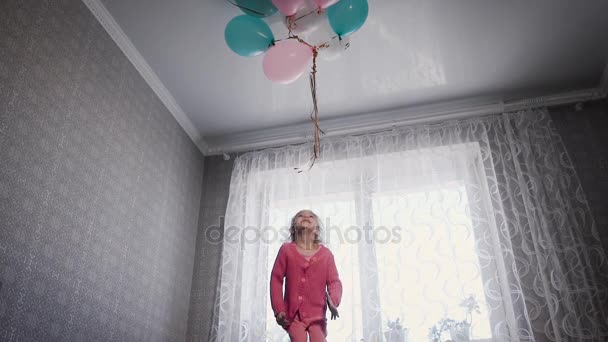 Niña feliz de pie cerca de la ventana en la cama en el vivero con el montón de globos de aire por encima. Pequeña niña está saltando en el sofá en la habitación tratando de atrapar bolas de colores . — Vídeo de stock