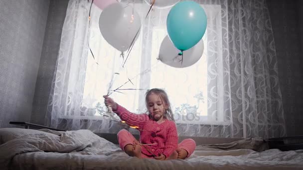 Nettes kleines Mädchen mit blonden Haaren, in rosa Pyjamas auf einem Bett der Eltern sitzend und mit Helium gefüllten Luftballons von Seite zu Seite winkend. die Kugeln in verschiedenen Farben - blau, weiß und rosa — Stockvideo