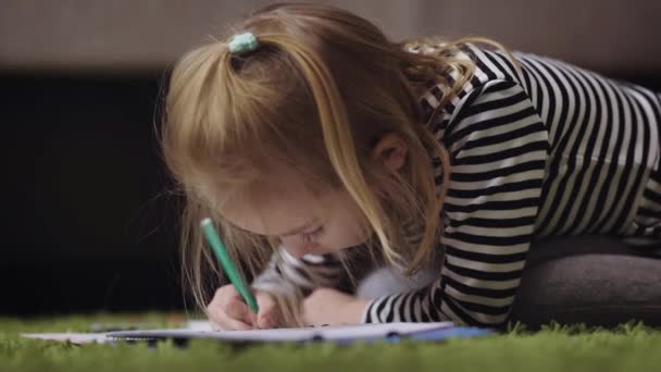 Het blonde meisje ligt op een tapijt in de kamer. De baby een groene viltstift tekent in een album op de schone lei. Op het meisje de gestreepte jas, grijze slipje, gele sokken. — Stockvideo