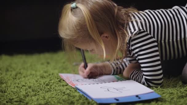 De baby ligt op een groene terry tapijt in de kamer. Het meisje een zwarte viltstift tekent in een notitieblok op de versierd bladen. Op het kind de gestreepte jas, grijze slipje, gele sokken. — Stockvideo