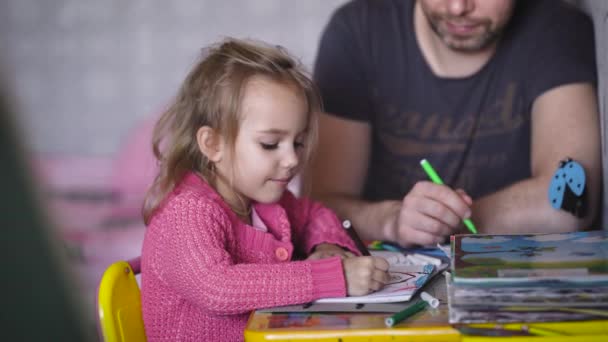 Malá dívka s plavými vlasy, oblečen v dětské halenku, je zapojena do výkresu spolu s jejím otcem. Malá holčička nakreslí dům a můj otec trávy. Oni jsou rádi, že spolu — Stock video