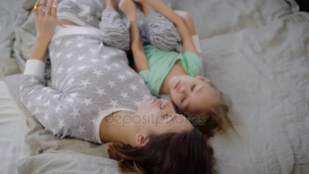 Мать с дочерью в спальне. Девушка с ребенком лежит на кровати и разговаривает. Ребенок в футболке мятного цвета и серых трусиках. Женщина в пижаме со звездами. Молодая семья . — стоковое видео