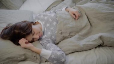 Genç esmer kadın portresi ile keten evde kapalı yalan ve yatakta uyuyan gri pijama giymiş. Güzel kız sabah, germe ve yeni güne gülümseyerek uyanıyor.