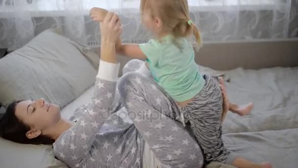 Mor med dotter i ett sovrum. Flickan med barnet ligga på en säng och spelas. Barnet i en t-shirt mint färg och grå trosor. Kvinnan i en pyjamas med stjärnor. Ung familj. — Stockvideo