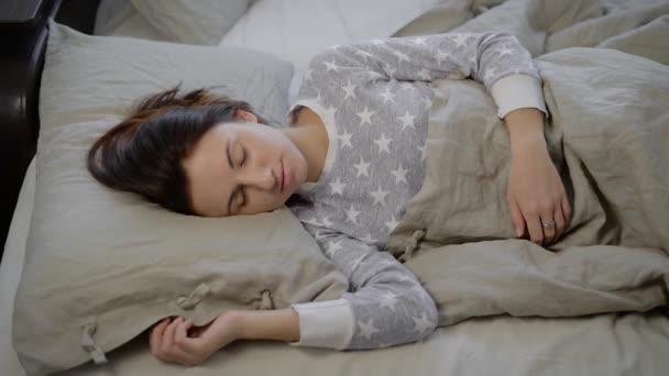 ベッドで女の子。若いブルネットは、午後に眠る。アスタリスクの付いたグレーのパジャマの女性。ブルネットのトスとターン。グレー色のベッドリネン. — ストック動画