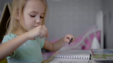 Sarı saçlı küçük şirin kız masa ve renkli Keçeli Kalemler berabere bir kağıt parçası üzerinde oturan mavi tişört giyiyor. Kız çizim, o bir sanatçı olurdu belki odaklı