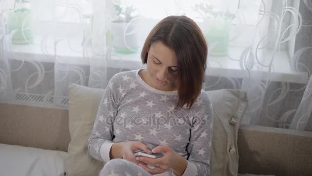 Flickan i en säng i ett sovrum. Unga brunett sitter på en säng. Kvinnan i en grå pyjamas med asterisker. Brunetten ser igenom nyheter på din smartphone. Sängkläder av grå färg. — Stockvideo