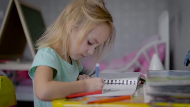 Het meisje in de kinderkamer aan een tafel. De baby wordt getekend in een schetsboek. In een hand bij het kind een viltstift kleur. Op een tafel van de pennen van het boek en vilt-tip van verschillende bloemen. — Stockvideo