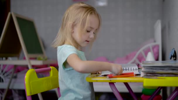 Маленька дівчинка малює картину з олівцями в студії, сидячи за столом. Маленька дитина вчиться малювати м'якими ручками різних кольорів. Мила дитина з задоволенням крепляє візерунок . — стокове відео
