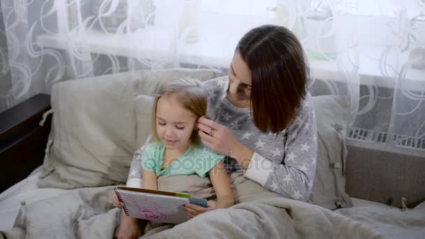 Une famille heureuse qui s'amuse au lit. Jolie jeune mère lit un livre à sa petite fille mignonne. Femme et petite fille sont assises ensemble, embrassant et discutant d'une queue de fée . — Video