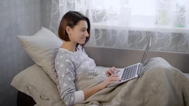 Het meisje op een bed in de kamer. De brunette semi-vergadering op een bed. De vrouw controleert de gegevens die op de laptop. Tegen de achtergrond van een venster met gordijnen. Beddengoed van grijze kleur. — Stockvideo