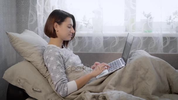 Portrait de belle femme brune couchée au lit avec ordinateur portable à la maison dans la chambre. Jeune fille avec une coupe de cheveux courte travaille comme pigiste passer sa journée de travail dans la chambre . — Video