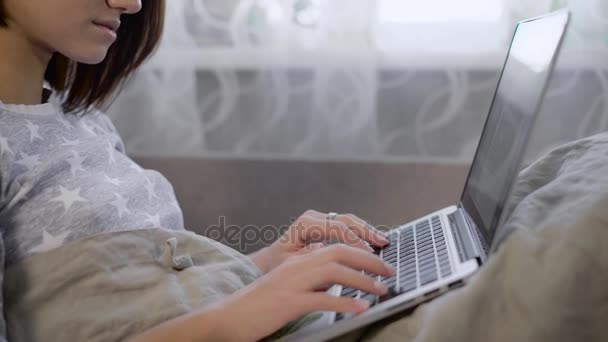 Młoda dziewczyna z brązowe włosy i stylowe włosów siedzi pod kołdrą i uważa, że odpowiedź na odebraną wiadomość od swojej przyjaciółki na laptopie. Ona spędza dni w łóżku. — Wideo stockowe