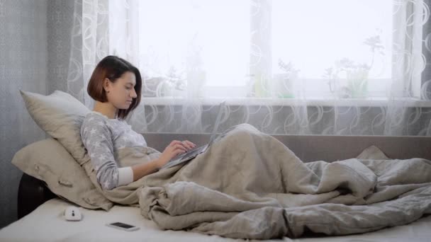Όμορφη μελαχρινή γυναίκα ντυμένη με piyamas ξαπλωμένη στο κρεβάτι με το laptop στο σπίτι στην κρεβατοκάμαρα. Νεαρή κοπέλα με το κοντό κούρεμα που εργάζεται ως ελεύθερος επαγγελματίας επαγγελματικές δαπάνες της εργάσιμης ημέρας στο δωμάτιο. — Αρχείο Βίντεο