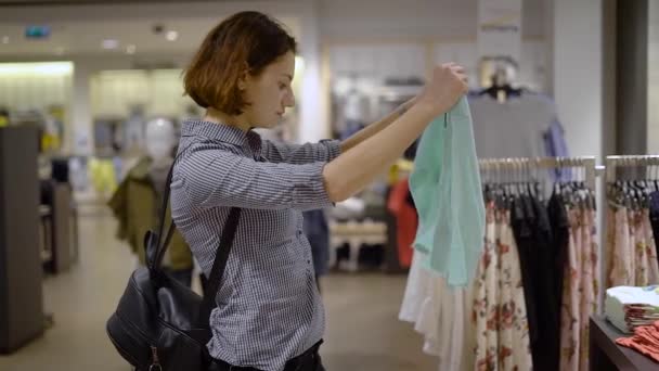 Een mooie, jonge meisje in een supermarkt kiest kleding. Het meisje ging in de boetiek te vinden van een nieuwe garderobe voor zichzelf. — Stockvideo
