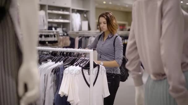 Una foto graciosa. Una chica en una tienda de ropa elige un atuendo. La mujer se sorprende por los precios caros, abraza una camisa o una blusa — Vídeo de stock