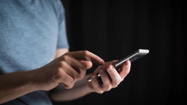 Conceito, vida online digital e redes sociais. Um homem de negócios em uma camiseta usa seu smartphone para ver suas contas. Tiro expressivo, um homem em um quarto escuro, segurando um telefone elegante — Vídeo de Stock