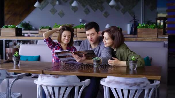 Tres jóvenes atractivos en un café o restaurante eligen un menú de bebidas y comida. Los amigos en el día libre vinieron a desayunar o almorzar . — Vídeos de Stock