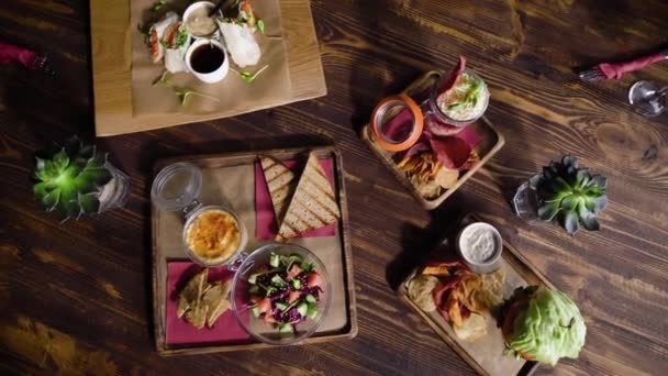 Gıda ile restoranda masa, en baştan görüntülemek. Güzel bir doku ile ahşap bir masa Snacks. Lezzetli sandviçler, soslar, salata ve patates kızartması. — Stok video