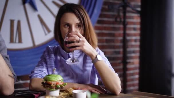 レストランで女の子は、ワインを飲んでいます。ブルネットは彼女のハンバーガーを持って、彼女はそれを食べる準備ができています。とても素敵な料理と軽食の提供 — ストック動画